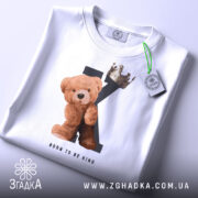 Футболка born king, білого кольору, складена на столі, фон світло-сірий, принт з ведмедиком та короною на літері K, текст 'BORN TO BE KING' під малюнком, на футболці є етикетка з зеленим шнурком.