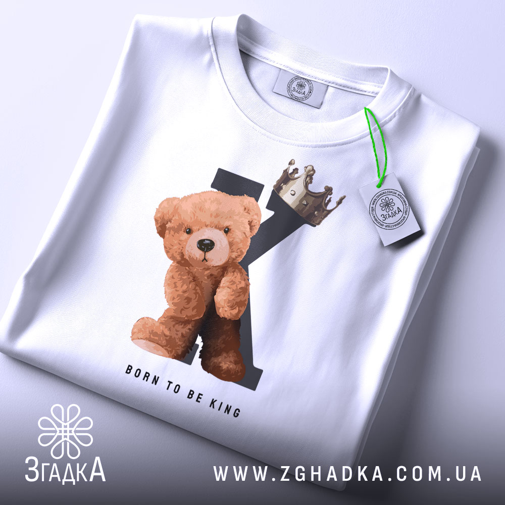 Футболка born king, білого кольору, складена на столі, фон світло-сірий, принт з ведмедиком та короною на літері K, текст 'BORN TO BE KING' під малюнком, на футболці є етикетка з зеленим шнурком.