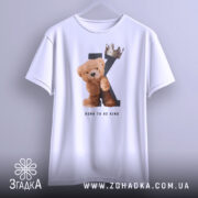 Футболка born king, білого кольору, підвішена на вішалці, фон світло-сірий, принт з ведмедиком та короною на літері K, текст 'BORN TO BE KING' під малюнком, рукави футболки вільні.