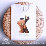 Футболка born king, білого кольору, складена на плетеній кошику, фон світло-сірий, принт з ведмедиком та короною на літері K, текст 'BORN TO BE KING' під малюнком, видно етикетку з написом.