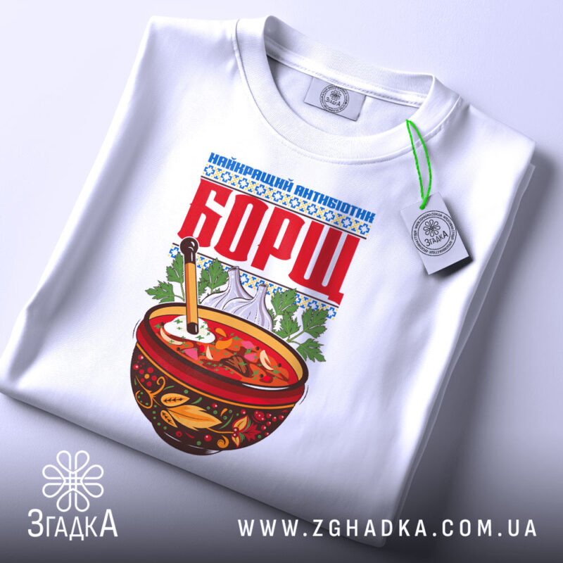 Футболка борщ подарунок, біла, складена на поверхні, фон світлий, принт з червоною написом 'БОРЩ' та малюнком тарілки з борщем, прикріплена етикетка.
