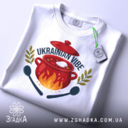 Футболка борщ український вайб, біла, складена на столі, фон світло-сірий, принт з червоним горщиком, вогнем, написом 'UKRAINIAN VIBE' та зеленими гілками, з етикеткою на зеленому шнурку.