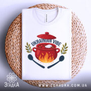 Футболка борщ український вайб, біла, складена на плетеній кошику, фон світло-сірий, принт з червоним горщиком, вогнем, написом 'UKRAINIAN VIBE' та зеленими гілками.