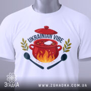 Футболка борщ український вайб, біла, складена на столі, фон світло-сірий, принт з червоним горщиком, вогнем, написом 'UKRAINIAN VIBE' та зеленими гілками, з етикеткою на зеленому шнурку.