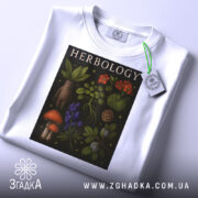 Футболка ботанічний арт, біла, складена на столі, фон світло-сірий, принт з рослинами та грибами, тег з логотипом прикріплений до коміра.