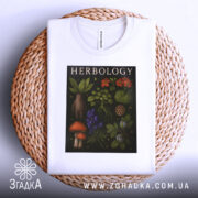 Футболка ботанічний арт, біла, складена на плетеній кошику, фон світло-сірий, принт з рослинами та грибами, текст 'HERBOLOGY' на чорному фоні.