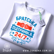 Футболка братська підтримка, біла, зображена зверху, на столі, фон світлий, принт яскравий з червоними та блакитними кольорами, напис 'Братська підтримка 24/7' та зображення кулаків, на футболці є етикетка.