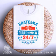 Футболка братська підтримка, біла, зображена зверху на плетеному кошику, фон світлий, принт яскравий з червоними та блакитними кольорами, напис 'Братська підтримка 24/7' та зображення кулаків.