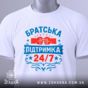 Футболка братська підтримка, біла, зображена зверху, на столі, фон світлий, принт яскравий з червоними та блакитними кольорами, напис 'Братська підтримка 24/7' та зображення кулаків, на футболці є етикетка.