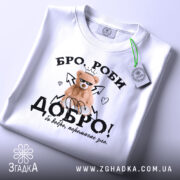 Футболка бро роби добро, біла, складена на столі, фон світлий, принт з ведмедиком у короні, напис 'ДОБРО!' з елементами сердець і стріл.
