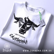 Футболка брутальна скотиняка, білого кольору, зображена зверху, на плоскій поверхні, фон світлий, принт чорного кольору з графічним зображенням голови корови та написом 'Брутальна скотиняка', з зеленим ярликом.