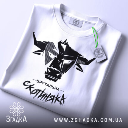 Футболка брутальна скотиняка, білого кольору, зображена зверху, на плоскій поверхні, фон світлий, принт чорного кольору з графічним зображенням голови корови та написом 'Брутальна скотиняка', з зеленим ярликом.