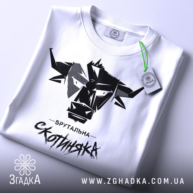 Футболка брутальна скотиняка, білого кольору, зображена зверху, на плоскій поверхні, фон світлий, принт чорного кольору з графічним зображенням голови корови та написом 'Брутальна скотиняка', з зеленим ярликом.