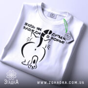 Футболка будь як дупця, білого кольору, складена на поверхні, фон світло-сірий, принт чорного кольору з малюнком кота ззаду та текстом, на комірці зелена етикетка.