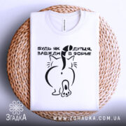 Футболка будь як дупця, білого кольору, складена на плетеній кошику, фон світло-сірий, принт чорного кольору з малюнком кота ззаду та текстом.