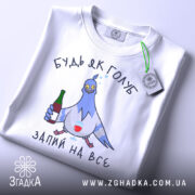 Футболка будь як голуб, білого кольору, зображена зверху, на ній видно принт з голубом, який тримає пляшку та келих, текст "ЗАПИЙ НА ВСЕ", на комірці є етикетка з зеленим шнурком.