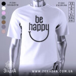 Футболка будь щасливим, білого кольору, зображена з близького ракурсу на манекені, фон світло-сірий, принт чорного кольору з написом 'be happy' і усмішкою.