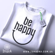 Футболка будь щасливим, білого кольору, складена на столі, фон світло-сірий, принт чорного кольору з написом 'be happy' та етикеткою з зеленим шнурком.