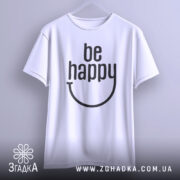 Футболка будь щасливим, білого кольору, підвішена на вішалці, фон світло-сірий, принт чорного кольору з написом 'be happy' і усмішкою.