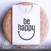Футболка будь щасливим, білого кольору, складена на плетеній кошику, фон світло-сірий, принт чорного кольору з написом 'be happy'.
