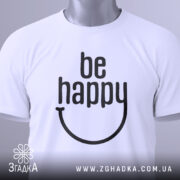 Футболка будь щасливим, білого кольору, складена на столі, фон світло-сірий, принт чорного кольору з написом 'be happy' та етикеткою з зеленим шнурком.