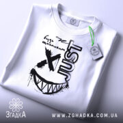 Футболка будь щасливим, біла, складена на столі, фон світло-сірий, принт чорного кольору з усмішкою та написом 'JUST', з зеленим ярликом.