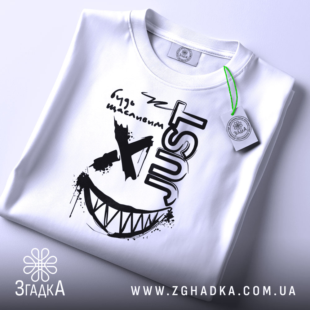 Футболка будь щасливим, біла, складена на столі, фон світло-сірий, принт чорного кольору з усмішкою та написом 'JUST', з зеленим ярликом.