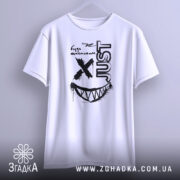 Футболка будь щасливим, біла, підвішена на вішалці, фон світло-сірий, принт чорного кольору з усмішкою та написом 'JUST'.