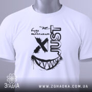 Футболка будь щасливим, біла, складена на столі, фон світло-сірий, принт чорного кольору з усмішкою та написом 'JUST', з зеленим ярликом.