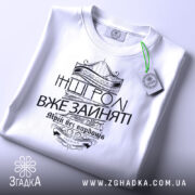 Футболка будь собою, біла, зображена з верхнього ракурсу, складена на поверхні, фон світло-сірий, принт чорного кольору з написом 'Будь собою' та 'Інші ролі вже зайняті', прикрашена етикеткою з зеленим шнурком.