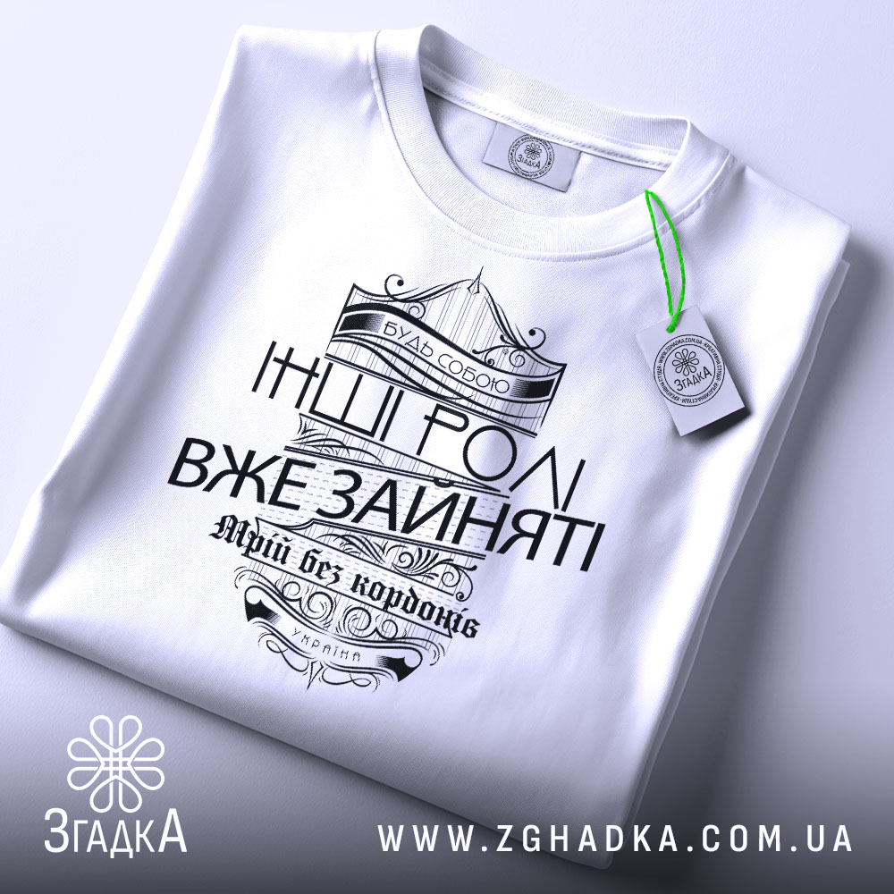 Футболка будь собою, біла, зображена з верхнього ракурсу, складена на поверхні, фон світло-сірий, принт чорного кольору з написом 'Будь собою' та 'Інші ролі вже зайняті', прикрашена етикеткою з зеленим шнурком.