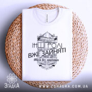 Футболка будь собою, біла, зображена з верхнього ракурсу, складена на плетеній кошику, фон світлий, принт чорного кольору з написом 'Будь собою' та 'Інші ролі вже зайняті', оформлений у стилі вензелів.