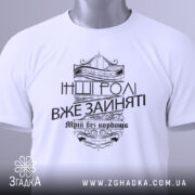 Футболка будь собою, біла, зображена з верхнього ракурсу, складена на поверхні, фон світло-сірий, принт чорного кольору з написом 'Будь собою' та 'Інші ролі вже зайняті', прикрашена етикеткою з зеленим шнурком.