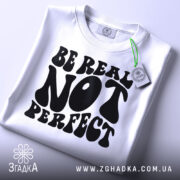 Футболка будь справжнім, білого кольору, складена на поверхні, з видимим етикеткою, фон світло-сірий, принт чорного кольору з написом 'BE REAL NOT PERFECT' у стилі хіпі.