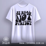 Футболка будь справжнім, білого кольору, підвішена на вішалці, фон світло-сірий, принт чорного кольору з написом 'BE REAL NOT PERFECT' у стилі хіпі.