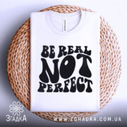 Футболка будь справжнім, білого кольору, складена на плетеній кошику, фон світло-сірий, принт чорного кольору з написом 'BE REAL NOT PERFECT' у стилі хіпі.