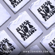 Футболка будь справжнім, білого кольору, зображена в фронтальному ракурсі на манекені, фон світло-сірий, принт чорного кольору з написом 'BE REAL NOT PERFECT' у стилі хіпі.