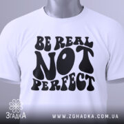 Футболка будь справжнім, білого кольору, складена на поверхні, з видимим етикеткою, фон світло-сірий, принт чорного кольору з написом 'BE REAL NOT PERFECT' у стилі хіпі.