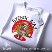 Футболка буенос діаз білого кольору, зображена на столі, фон світлий, принт з собакою в капелюсі та червоному шарфі, на футболці є етикетка.