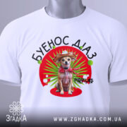 Футболка буенос діаз білого кольору, зображена на столі, фон світлий, принт з собакою в капелюсі та червоному шарфі, на футболці є етикетка.