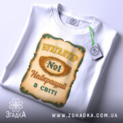 Футболка бухгалтер номер один, біла, зображена зверху, фон світлий, принт з написом 'БУХГАЛТЕР No1 Найкращий в світі' з елементами декору, на футболці є етикетка.