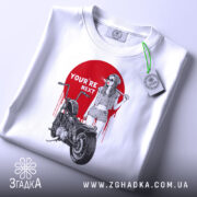 Футболка бунтарський настрій, біла, складена на поверхні, фон світло-сірий, принт з червоним колом, дівчина в окулярах з бейсбольною битою, мотоцикл поруч, текст 'YOU'RE NEXT' на принті, з зеленим ярликом.