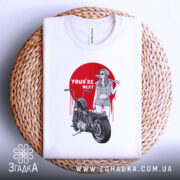 Футболка бунтарський настрій, біла, складена на кошику, фон світло-сірий, принт з червоним колом, дівчина в окулярах з бейсбольною битою, мотоцикл поруч, текст 'YOU'RE NEXT' на принті.