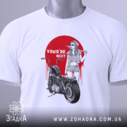Футболка бунтарський настрій, біла, складена на поверхні, фон світло-сірий, принт з червоним колом, дівчина в окулярах з бейсбольною битою, мотоцикл поруч, текст 'YOU'RE NEXT' на принті, з зеленим ярликом.