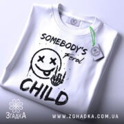 Футболка бунтівна дитина, біла, зображена зверху, на ній видно етикетку з зеленим шнурком, фон світлий, принт аналогічний попередньому, текст та малюнок виконані в чорному кольорі.