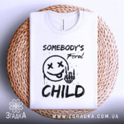 Футболка бунтівна дитина, біла, зображена зверху на плетеній основі, фон світлий, принт з смайлом та текстом 'SOMEBODY'S Feral CHILD' у чорному кольорі, видно деталі тканини.