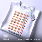 Футболка бути іншим. Біла футболка, складена на поверхні, з принтом оранжевих автомобілів і зеленим велосипедом. На футболці є етикетка з зеленим шнурком. Фон світлий, а ракурс зверху.