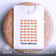 Футболка бути іншим. Біла футболка, складена на плетеній кошику, з принтом оранжевих автомобілів і зеленим велосипедом. Фон світлий, а ракурс зверху.