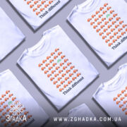 Футболка бути іншим. Біла футболка з принтом, на якому розташовані оранжеві автомобілі в ряд, а один зелений велосипед виділяється серед них. Фон нейтральний, а ракурс зосереджений на передній частині футболки.