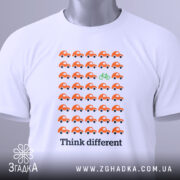 Футболка бути іншим. Біла футболка, складена на поверхні, з принтом оранжевих автомобілів і зеленим велосипедом. На футболці є етикетка з зеленим шнурком. Фон світлий, а ракурс зверху.